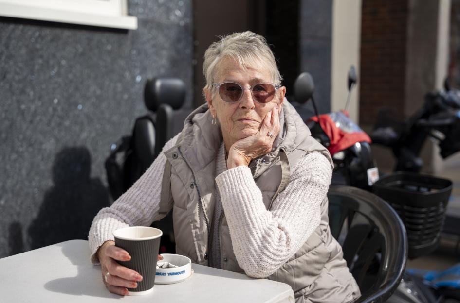 An older woman with short gray hair and sunglasses, wearing a vest over a sweater, sits at a table outdoors, holding a coffee cup and resting her chin on her hand.