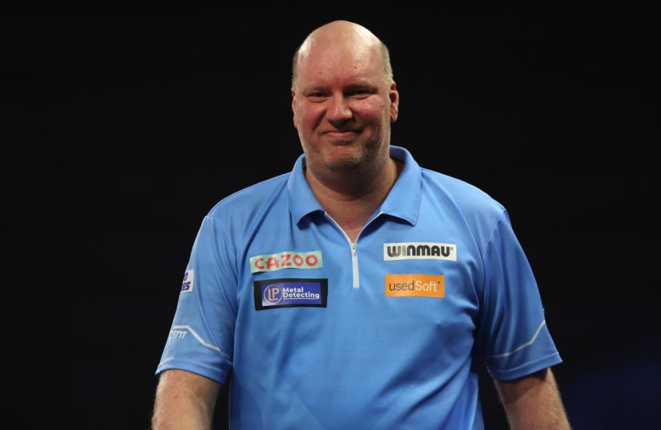 A male darts player in a light blue shirt with several sponsors smiles at the camera.