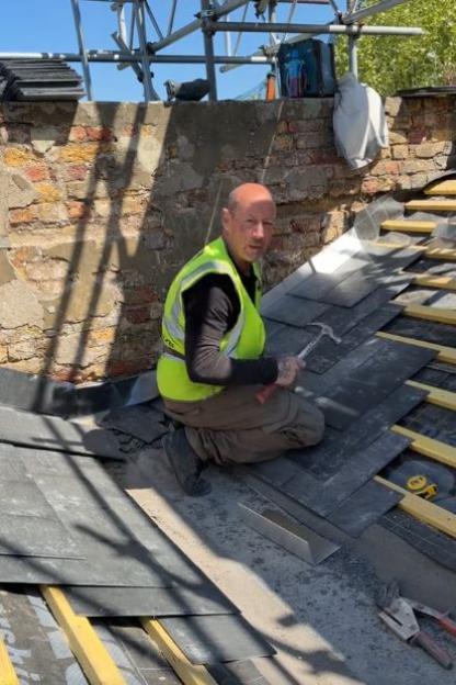A man in a safety vest kneeling on a roof with a hammer in his hand.