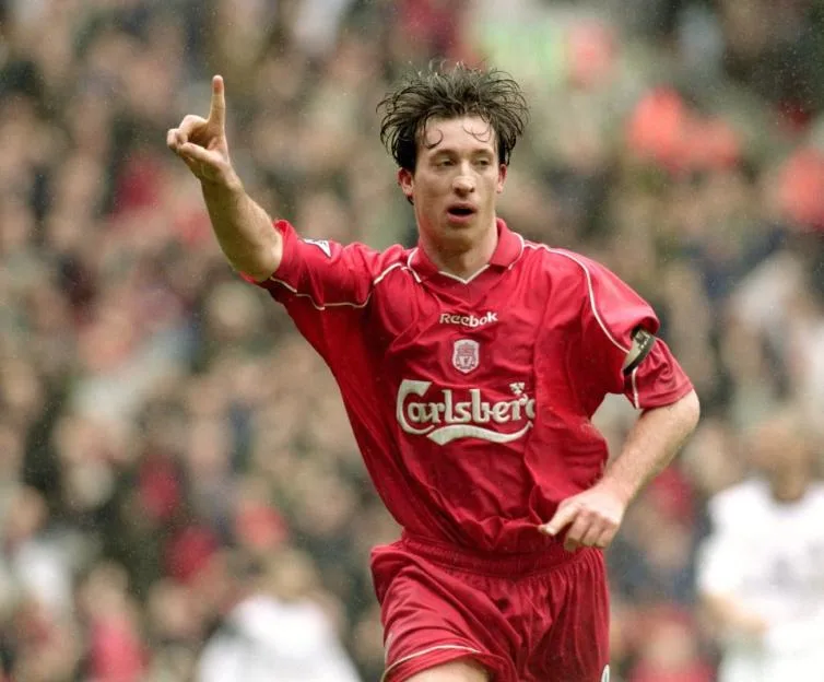 Robbie Fowler in a red Liverpool uniform celebrates a goal by pointing his index finger upwards.