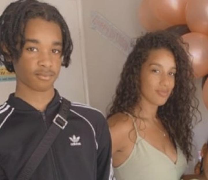 A woman and two children stand indoors in front of birthday balloons.