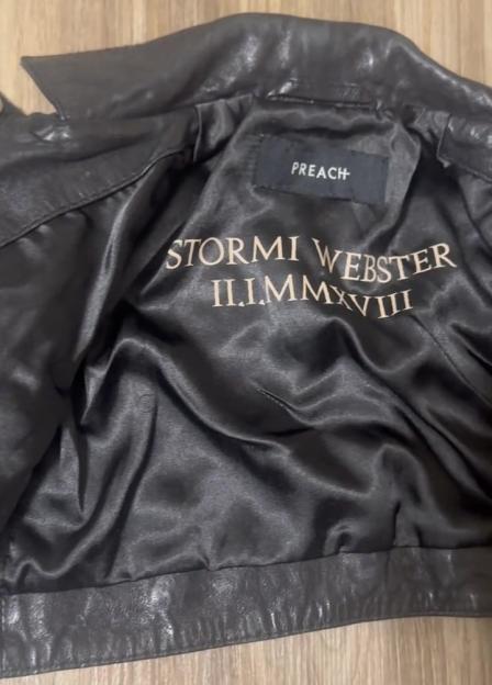 The inner lining of a black leather jacket with "STORMI WEBSTER" and "II.I.MMXVIII" embroidered in gold.