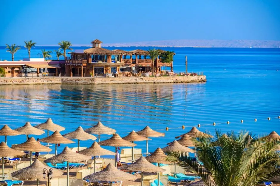 A beach resort in Hurghada, Egypt, featuring straw parasols, lounge chairs, and a seaside building under a clear blue sky.