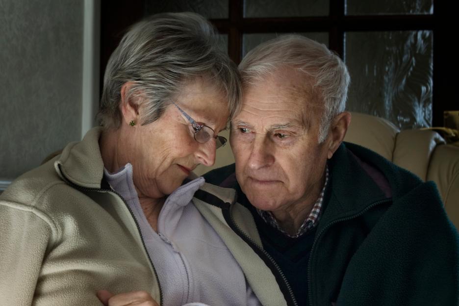 Elderly couple wearing coats indoors, appearing cold.