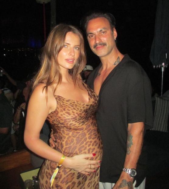 A man with a mustache and a woman in a leopard print dress, with her hand on her pregnant belly, pose together.