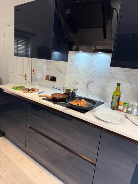 A newly renovated kitchen with dark blue cabinets, marble backsplash, cooking pans on an induction stove, and ingredients for a meal.