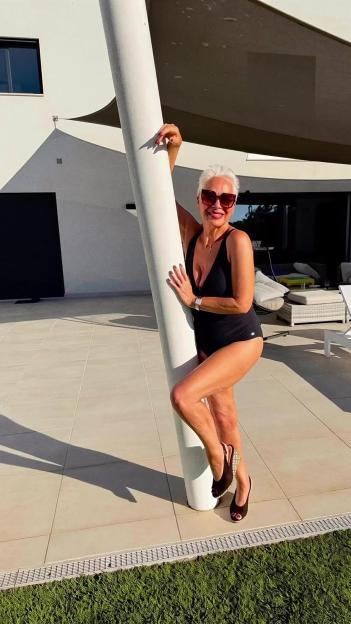 Denise Welch poses in a black swimsuit and heels against a white column by a pool.