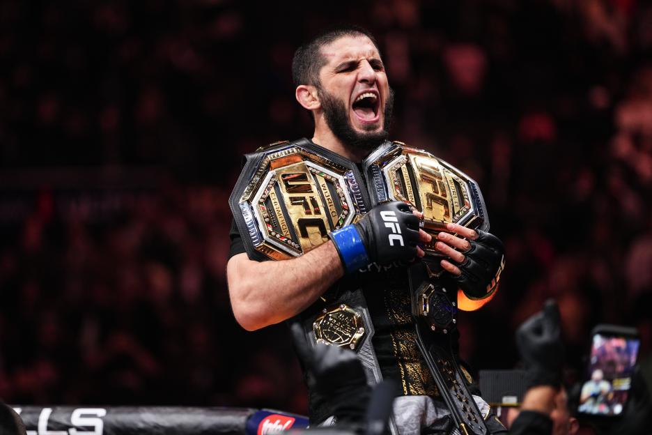 Islam Makhachev reacts to his win, wearing multiple UFC championship belts.