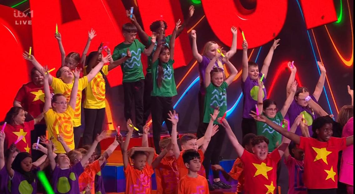 Children on stage in brightly colored t-shirts holding glowing sticks with arms raised.