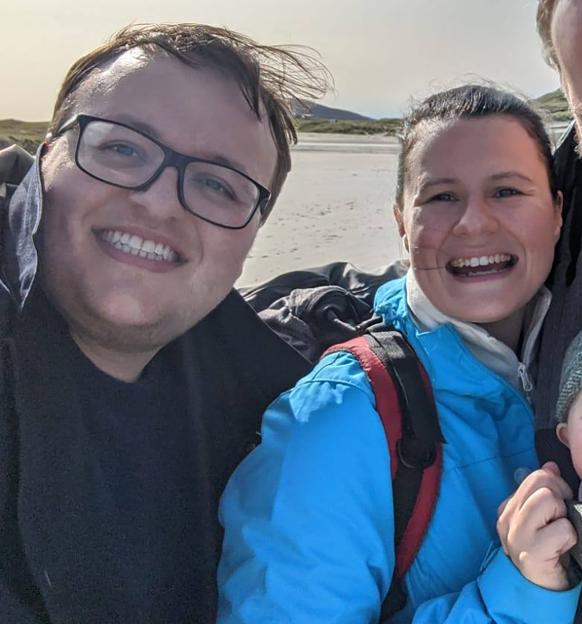 David Taylor and Joani Reid smiling at the camera with a beach and hills in the background.