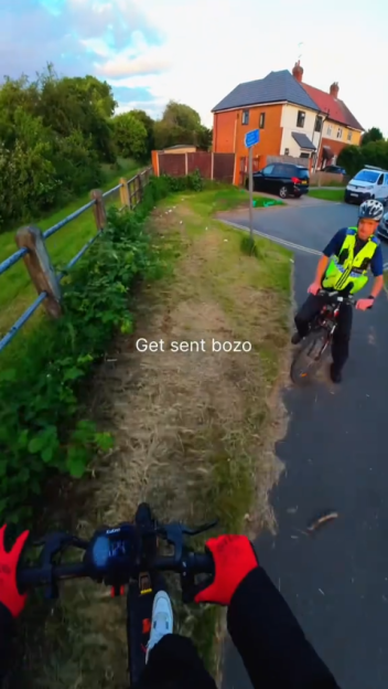 Point-of-view shot from an e-scooter, with the driver's gloved hands on the handlebars, showing a police officer on a bicycle to the right.
