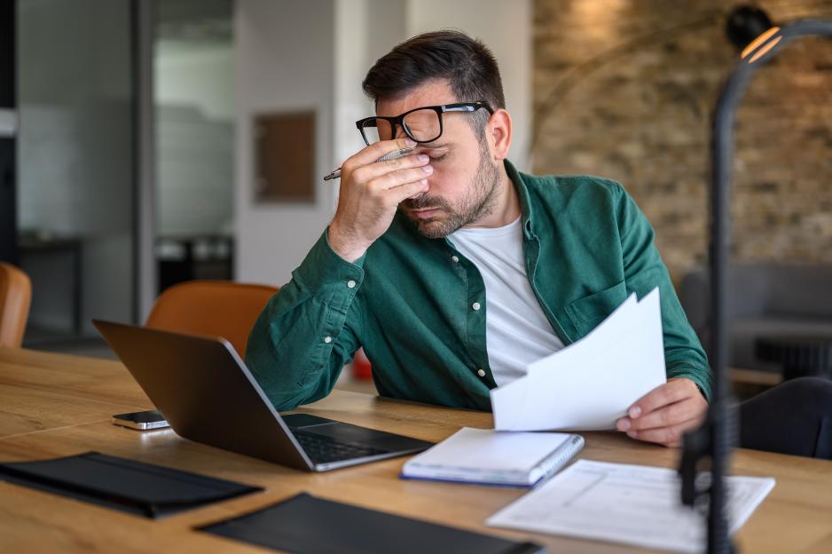 Anxious man rubs his eyes while working at a desk with a laptop and papers.