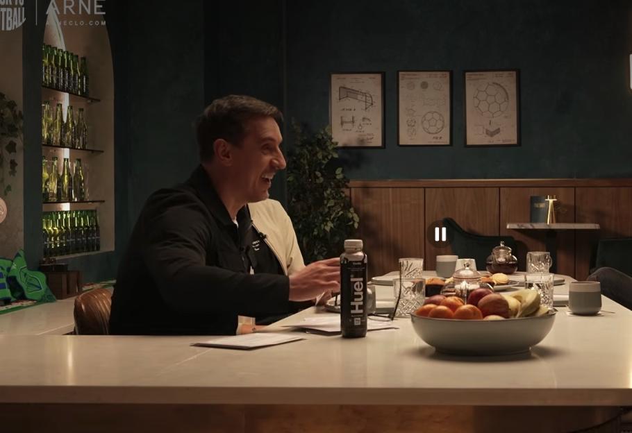 A man with short dark hair in a black jacket sits at a table, smiling while looking to the right, with a bowl of fruit and a bottle labeled "Huel" in front of him.