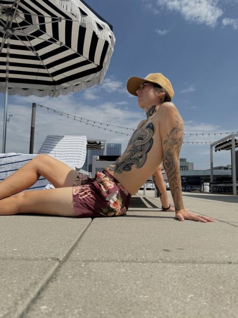 A person with a tattooed torso and arms, wearing floral swim trunks and a cap, sits on a paved surface next to a striped umbrella.