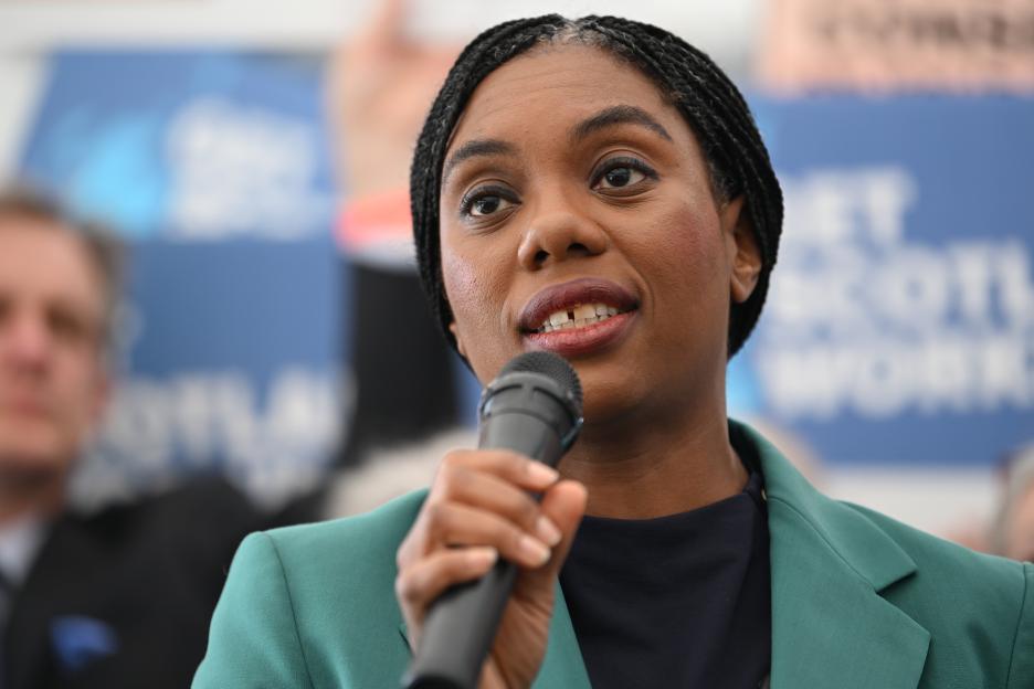 A woman with braided hair and a teal blazer speaks into a microphone.