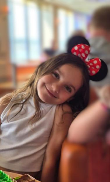 Aria Thorpe, a young girl with long brown hair, smiles while wearing Minnie Mouse ears.