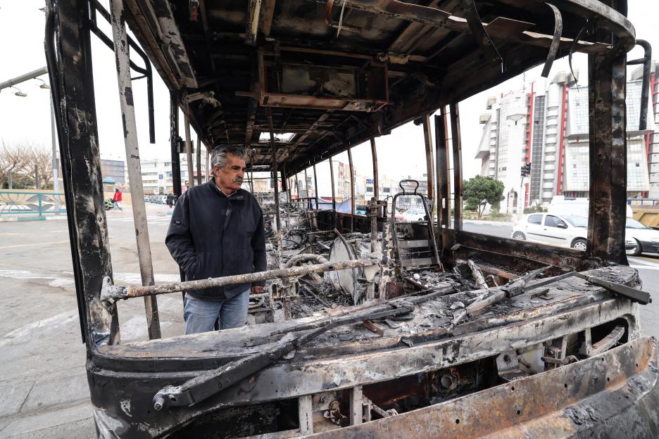 A man stands inside a burnt bus with a banner about public funds on display in Sadeghieh Square, Tehran, Iran.