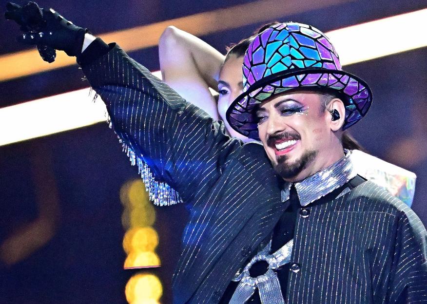 British musician Boy George performing in a shiny black pinstriped suit, with a rainbow-colored patterned top hat, and glittery makeup.