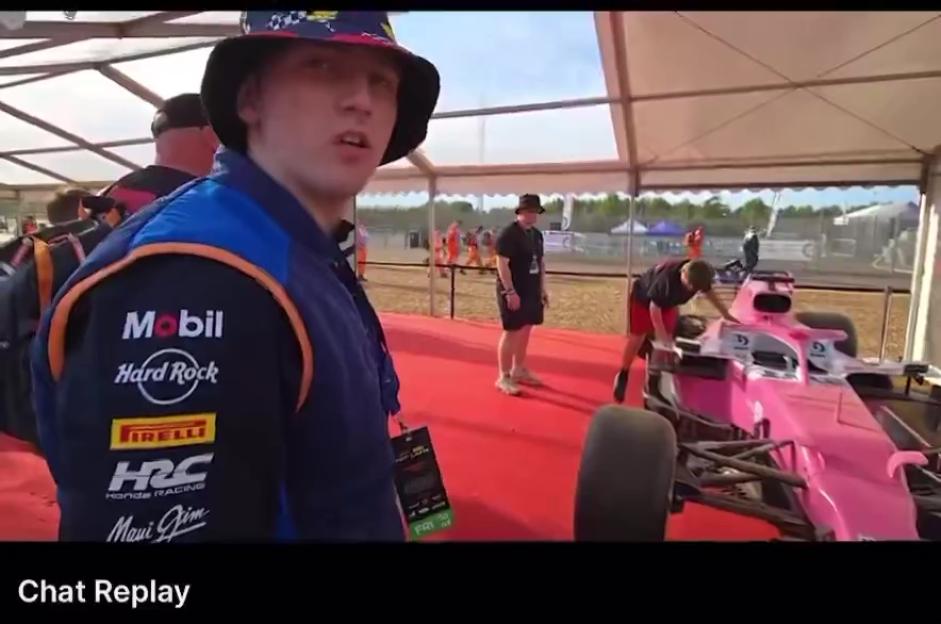 A young man in a bucket hat and racing jacket stands in front of a pink race car under a tent.