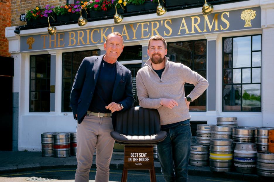 Two men stand outside "The Bricklayer's Arms" pub with a branded chair that reads "Best Seat In The House".