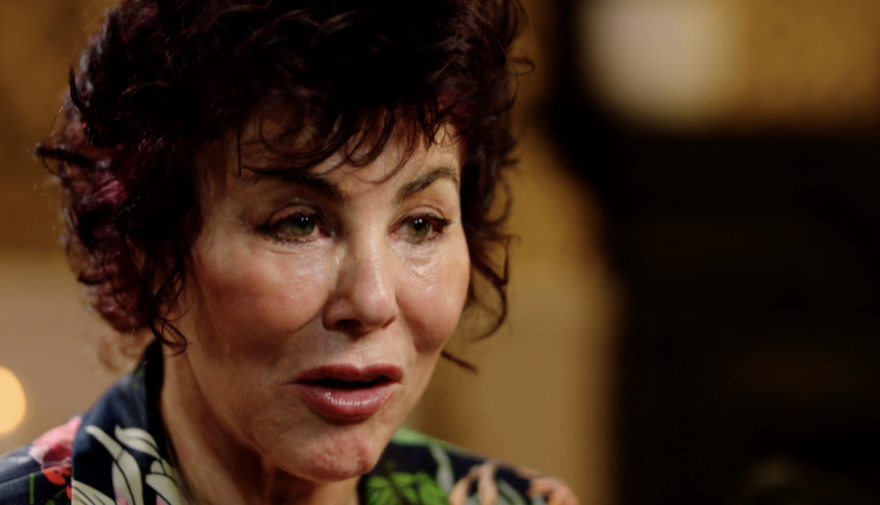 A close-up of a woman with short, curly dark hair and green eyes, wearing a floral shirt, speaking.