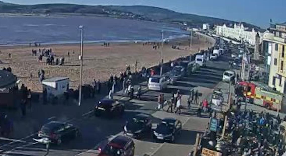 Overhead shot of a beach and an adjacent road with people, vehicles, and an emergency response truck.