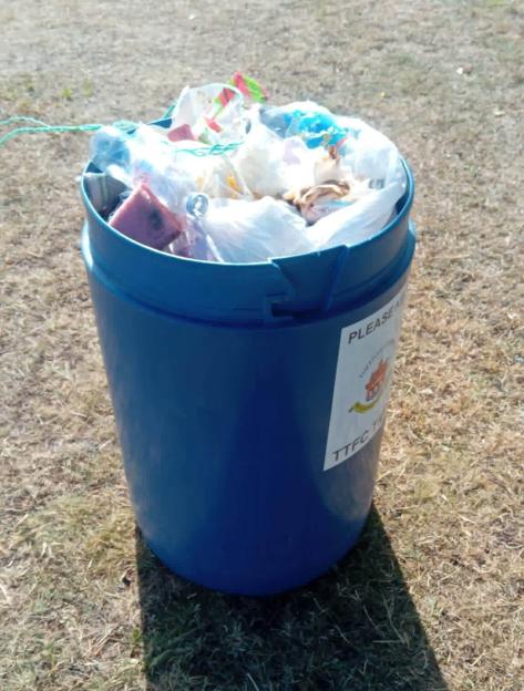 A blue rubbish bin overflowing with trash, including plastic bags and food waste, sitting on a patch of dry grass.