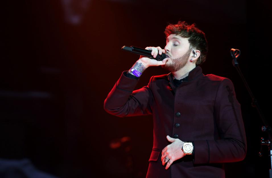 James Arthur performing on stage, wearing a dark red coat and holding a microphone to his mouth.