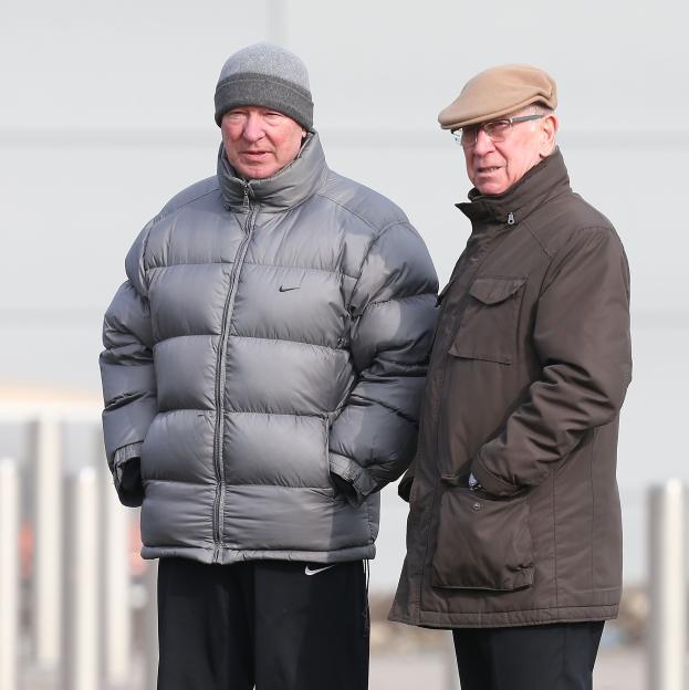 Alex Ferguson in a grey puffer jacket and beanie, and Bobby Charlton in a brown jacket and flat cap, looking on.