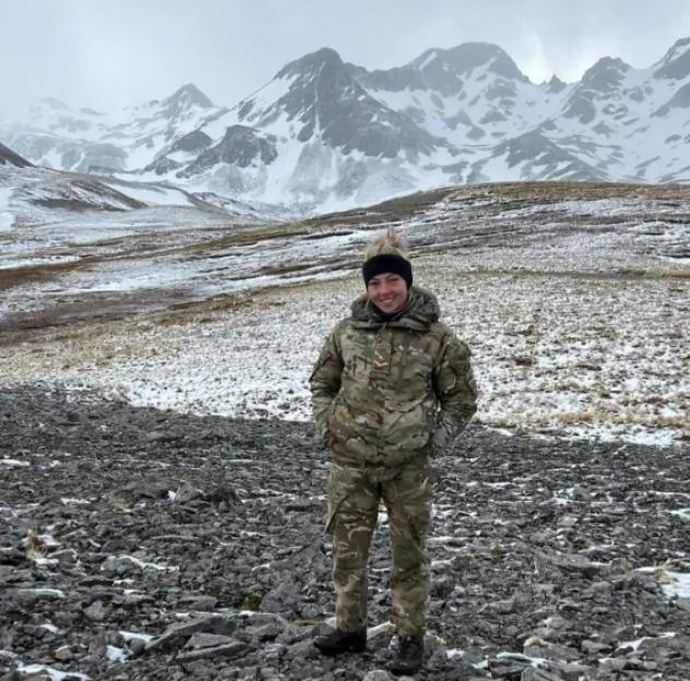 A soldier in camouflage and a black beanie stands in a rocky, snowy landscape with mountains in the background.