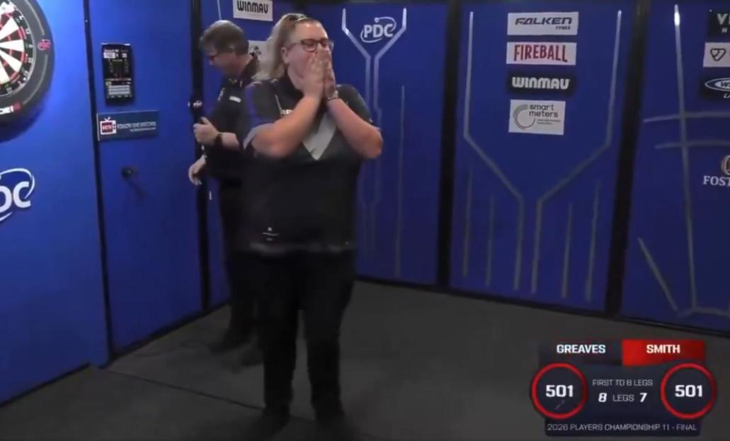 A female darts player with blonde hair in a ponytail covers her mouth with both hands in surprise, while another player stands behind her. In the foreground, a scoreboard shows "GREAVES" with a score of 501 and "SMITH" with a score of 501. It also displays "FIRST TO 8 LEGS" with "8 LEGS 7" and "2026 PLAYERS CHAMPIONSHIP 11 - FINAL".
