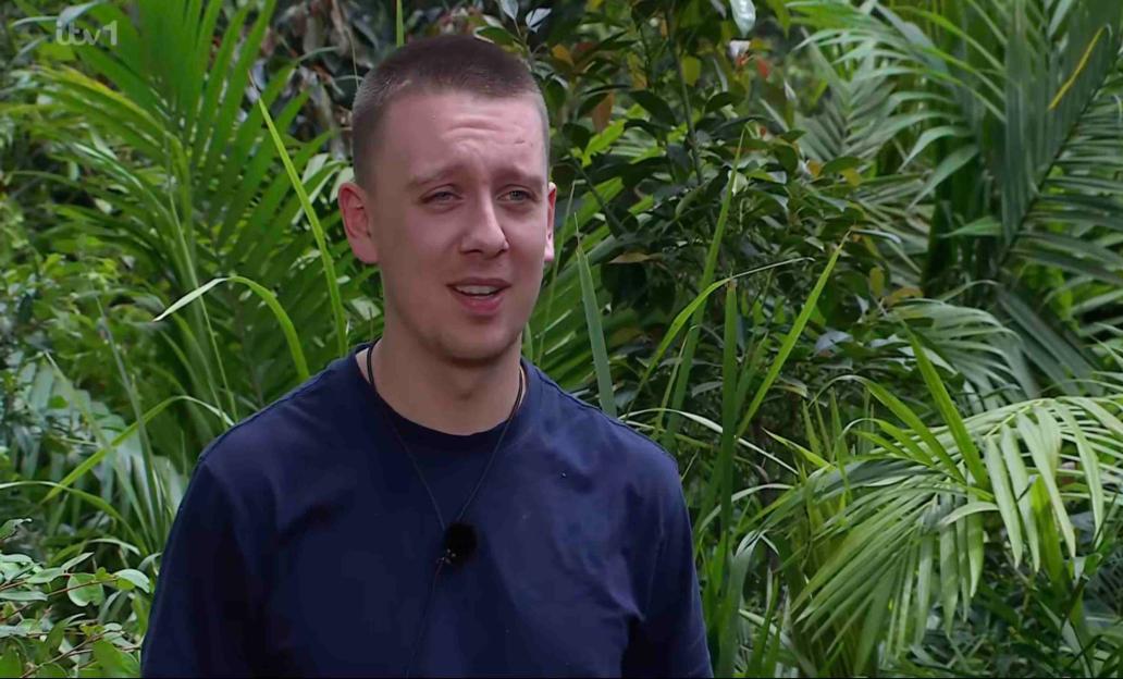 Aitch in a dark blue shirt with a microphone, against a background of lush green jungle foliage.