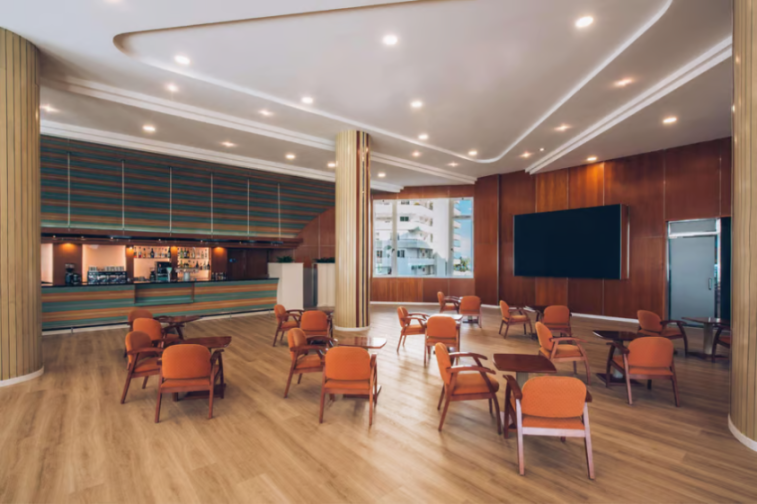 A lounge area with a long bar, orange chairs, and a large television on the wall.
