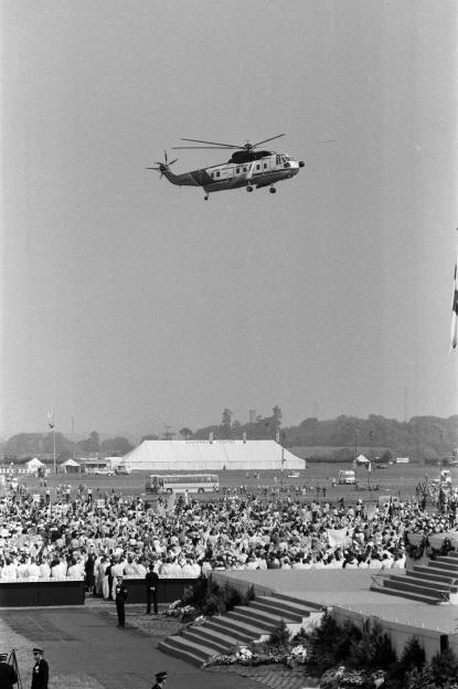Black and white photo of a helicopter flying over a crowd of people gathered for a religious mass at Coventry Airport.