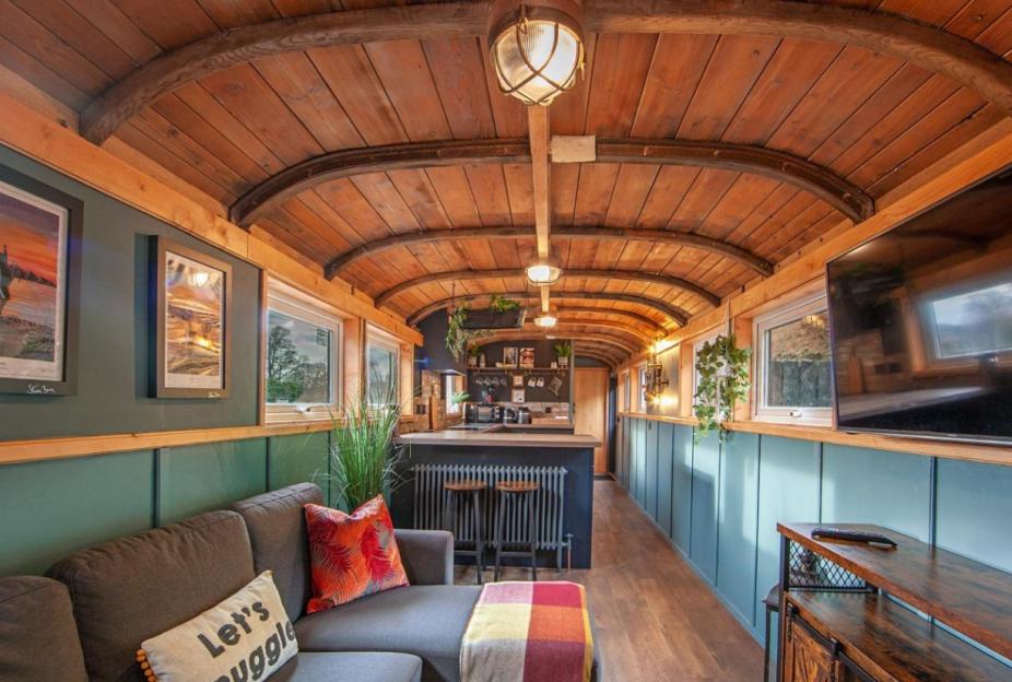 A narrowboat interior with a wooden arched ceiling, a grey sectional sofa, and a kitchen with bar stools.