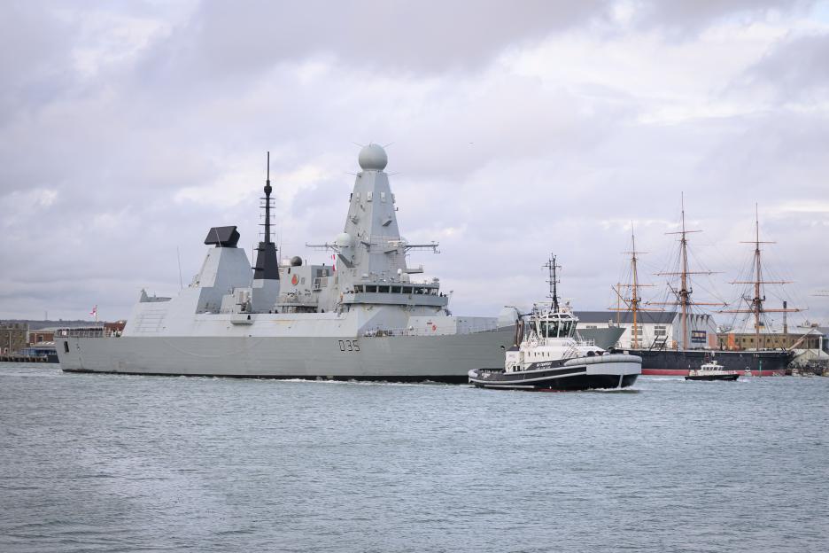 The destroyer HMS Dragon (D35) sets sail from Portsmouth Harbour, escorted by a tugboat and with a historic sailing ship docked in the background.