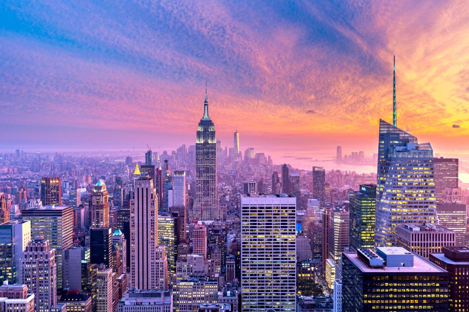 A high-angle view of the Manhattan skyline at sunset, with the Empire State Building and other skyscrapers brightly lit against a purple, pink, and orange sky.