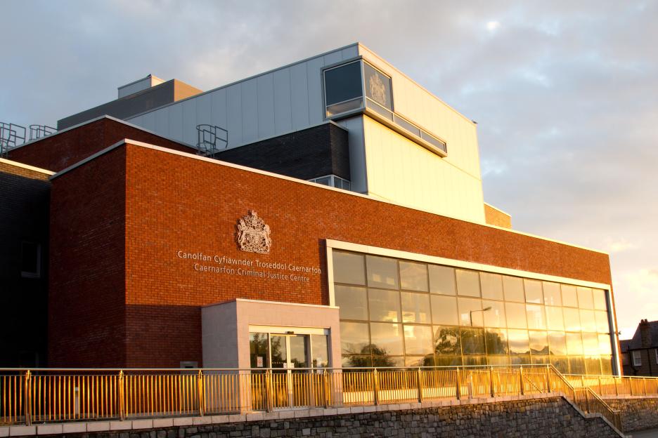 The Caernarfon Criminal Justice Centre building with "Canolfan Cyfiawnder Troseddol Caernarfon" and the Royal Coat of Arms on the brick facade.