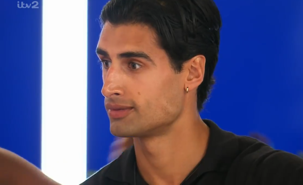 Samraj, a man with dark hair, a hoop earring, and a black shirt, looks to the right against a blue and white background.