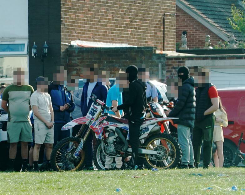 A group of people, with faces blurred, gathered outdoors, with two dirt bikes on the grass in the foreground and a brick building in the background.