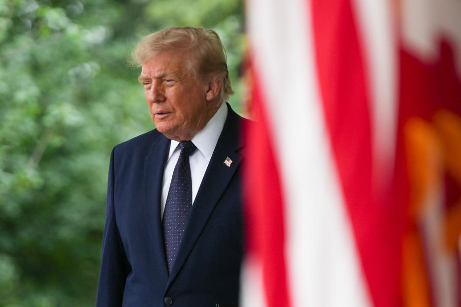 President Donald Trump in a dark suit and tie with an American flag pin, standing outdoors with a blurred green background.