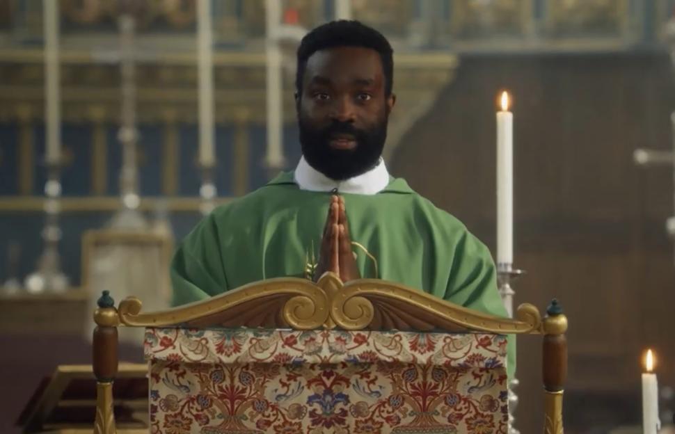 A man in a green robe stands behind an ornate lectern, hands clasped, in a church.