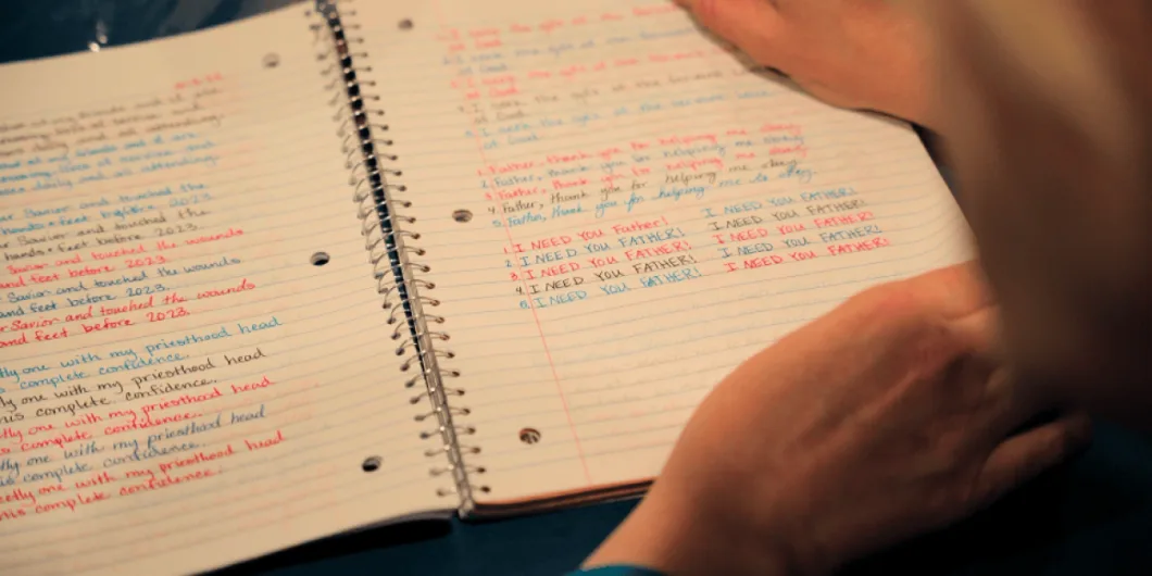 A close-up of a person holding an open spiral-bound notebook with handwritten prayers and phrases in red and blue ink, including "I NEED YOU FATHER!".