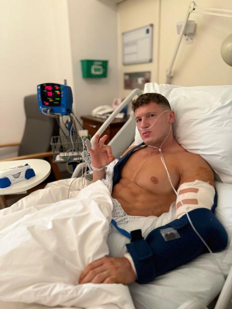 A man with a bandaged arm and chest, a nasal cannula, and a heart rate monitor making an "OK" sign in a hospital bed.