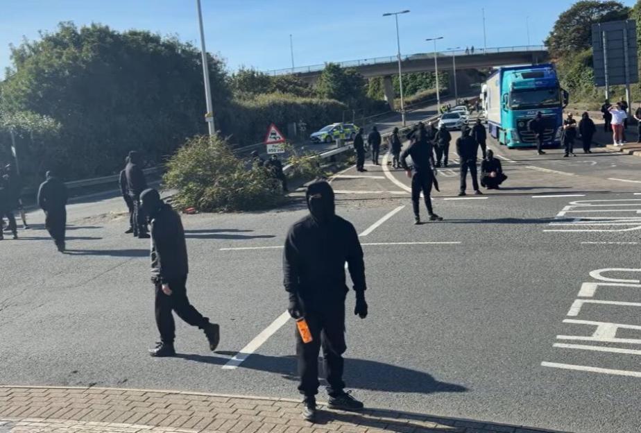 Protestors in black clothing and face coverings blocking a road with a police car and truck visible in the background.