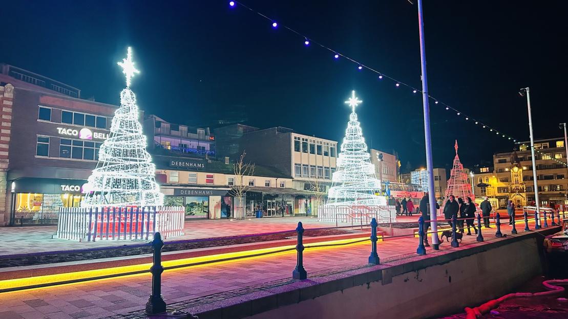 Nighttime view of a street decorated for Christmas with two large, illuminated Christmas tree structures and people walking along the sidewalk.