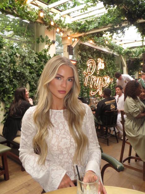 Olivia Livvy Barker from Made In Chelsea in a white lace top, sitting at a table with a drink, in a restaurant decorated with plants and string lights.