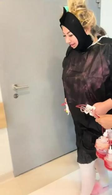 A woman walking in a hospital gown and face wrap, carrying multiple fluid drainage bulbs.