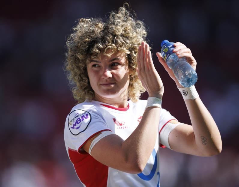 A female rugby player in a white jersey claps while holding a water bottle during the Women's Guinness Six Nations.