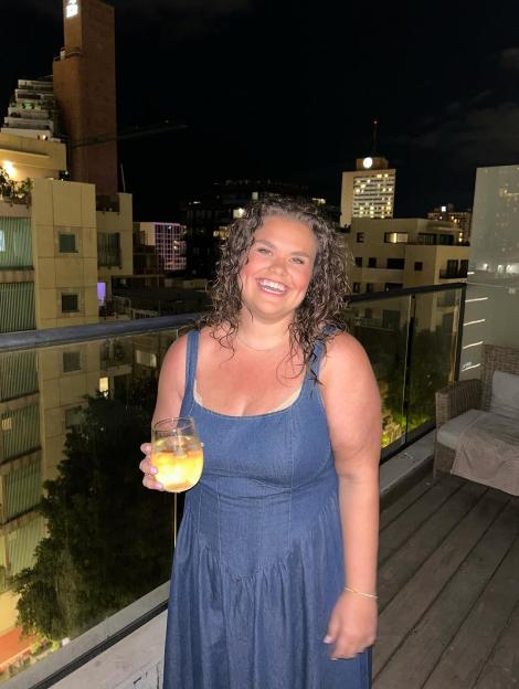 Amy Tapper in a denim dress holding a drink on a balcony at night.